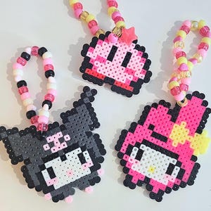 Puede incluir: Tres collares de cuentas de colores con colgantes con dijes. Los dijes son diseños pixelados de personajes de dibujos animados en rosa, negro y blanco. Las cuentas son rosas, amarillas y blancas. Los collares están sobre una superficie blanca.