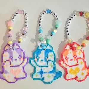 Puede incluir: Tres llaveros de cuentas coloridos con diseños de perros pixelados. Cada perro es de un color diferente: morado, azul y naranja. Los llaveros tienen bucles de cuentas con las palabras "AWAY" y "SANCTUARY".