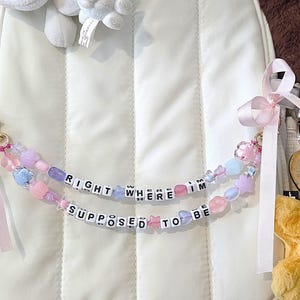 Puede incluir: Una mochila blanca con una correa de cuentas que dice "RIGHT WHERE I'M SUPPOSED TO BE". Una etiqueta de madera en forma de corazón con el texto "Never forget the difference you make" cuelga de la mochila. Se ven juguetes de peluche.