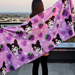 May include: A lavender scarf featuring a repeating pattern of a cartoon character with a black and white face, strawberries, skulls, hearts, and stars. The scarf is held up against a cityscape background.