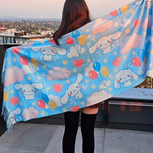 May include: A light blue scarf featuring a repeating pattern of white cartoon bunnies, clouds, balloons, and hearts. The scarf has a fringed edge and is held up against a cityscape background.