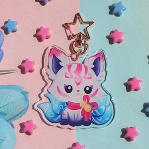 May include: A clear acrylic charm featuring a cartoon fox with white, pink, and blue details. The charm has a gold star-shaped clasp and is surrounded by pink, blue, and purple star-shaped embellishments. The fox has a pink bow.