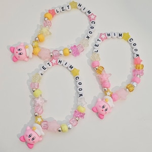 Puede incluir: Tres pulseras de cuentas con el texto "LET HIM COOK". Las pulseras presentan cuentas rosas, amarillas y transparentes, detalles en forma de estrella y un dije de Kirby rosa. Las pulseras están sobre una superficie blanca.