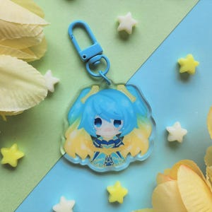 May include: A blue anime-style character keychain with a blue clasp. The character has blue hair and a yellow and blue outfit. The background is a mix of light green and blue, with yellow and white star-shaped decorations and yellow flower petals.