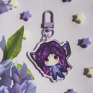 May include: A purple and blue acrylic charm featuring a cartoon character with purple hair and a star-shaped design. The charm is attached to a purple clasp. The background includes purple and yellow star-shaped decorations and purple flowers.