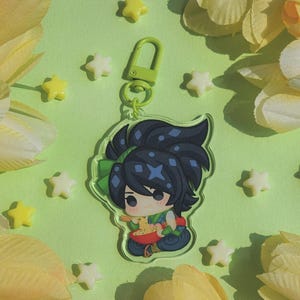 May include: A clear acrylic keychain featuring a cartoon character with dark blue hair and a green bow. The character is holding a bowl of food. The keychain has a green clasp and is surrounded by yellow and white star-shaped decorations.