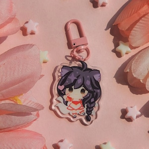 May include: A clear acrylic charm featuring a cartoon character with purple hair and a red outfit, attached to a pink clasp. The charm is surrounded by pink flower petals and star-shaped decorations on a pink background.