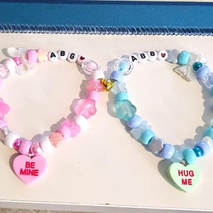 Puede incluir: Dos pulseras de cuentas. Una es rosa y blanca con un colgante en forma de corazón que dice "BE MINE". La otra es azul y blanca con un colgante en forma de corazón que dice "HUG ME". Ambas pulseras tienen los nombres "ABG" y "ABB" escritos con cuentas.