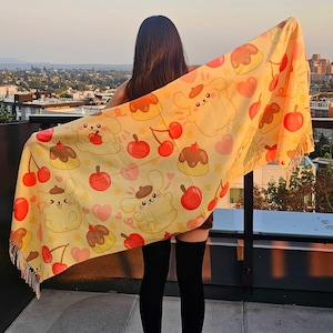 May include: A yellow scarf with a repeating pattern of cartoon characters, cherries, and desserts. The scarf has a fringe along the bottom edge. The background shows a cityscape under a sunset.