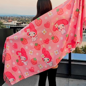 May include: Pink scarf featuring a repeating pattern of a cartoon character with a flower on its head, strawberries, hearts, and flowers. The scarf has a fringed edge. The background is a cityscape.