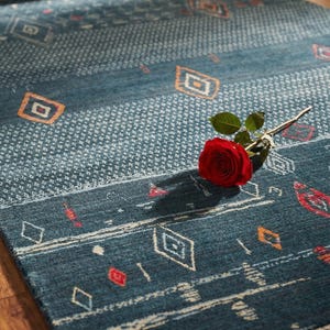 May include: A blue rectangular rug with geometric patterns in white, orange, and red. A red rose rests on the rug. The rug has a fringed edge and is on a wooden floor. The rug's dimensions are in centimetres.