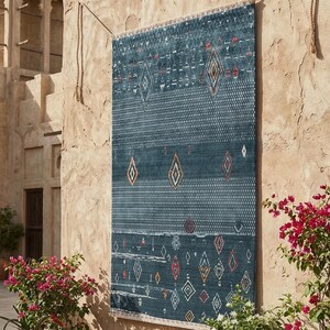 May include: A teal-coloured rectangular rug with a geometric diamond and triangle pattern. The rug is displayed on a beige wall, with a fringe along the bottom edge. The design incorporates orange, red, and white accents.