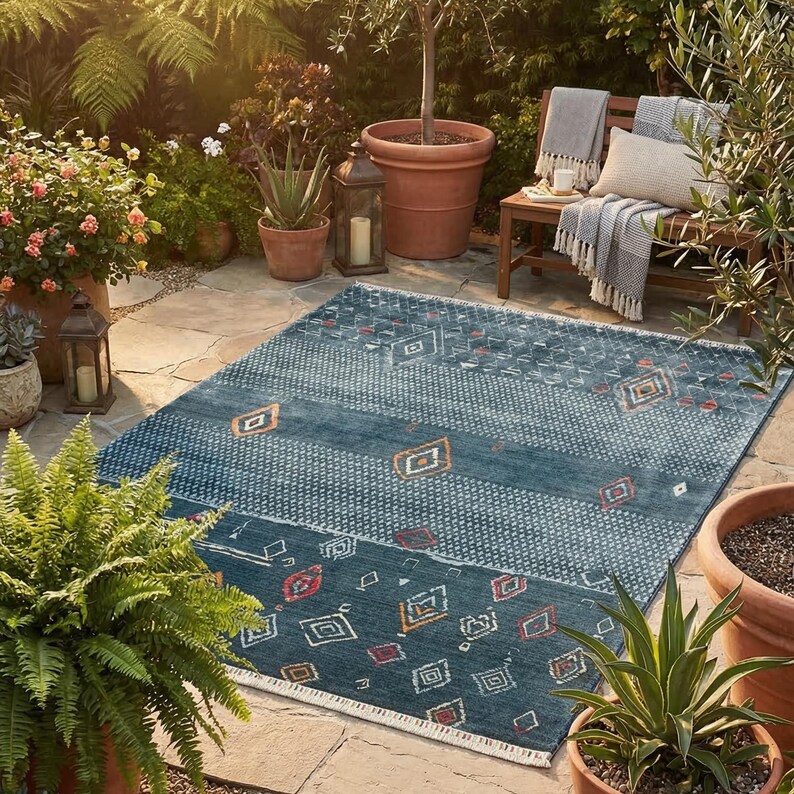 May include: A rectangular blue outdoor rug with geometric patterns in white, orange, and red. The rug is placed on a stone patio, surrounded by potted plants, a wooden bench, and lanterns, creating a cosy outdoor living space.