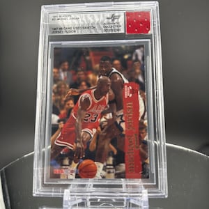 May include: A basketball trading card featuring Michael Jordan in a red Chicago Bulls jersey, dribbling a basketball. The card is encased in a protective holder. Text includes "1995-96 HOOPS #21 MICHAEL JORDAN" and "1987-88 GAME USED SWATCH JERSEY FUSION."