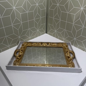 May include: A rectangular gold-colored decorative tray with ornate detailing and handles. The tray has a mirrored surface and is presented in a white box. The tray's design features intricate scrollwork and floral accents.
