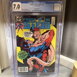 May include: A graded comic book titled "Power of the Atom #14" from DC Comics, featuring a vibrant illustration of two costumed figures in a fight scene. The cover has a yellow background with the text "HUMBUG'S BACK AND YOU'RE GONNA BE IN TROUBLE..."
