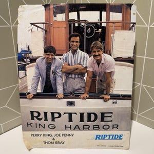 May include: A vintage Riptide King Harbor poster featuring Perry King, Joe Penny, and Thom Bray. The poster shows the three men on a boat. The Riptide logo is in blue. The poster is slightly worn.
