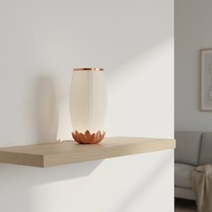 Può includere: Una lampada da tavolo con paralume cilindrico in tessuto bianco sporco e base color rame a forma di fiore di loto. La lampada presenta un bordo coordinato color rame sulla parte superiore ed emette una luce calda e soffusa.