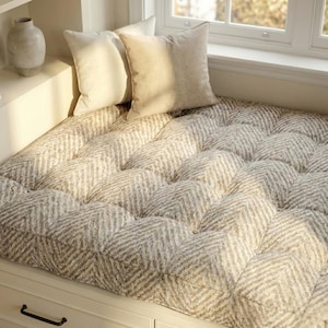 May include: A window seat with a tufted cushion in a neutral herringbone pattern. Two square pillows in a solid, light beige fabric rest against the window. A decorative vase sits on a nearby shelf.