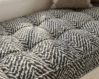 May include: A tufted bench cushion with a black and white herringbone pattern. The cushion is rectangular and has a series of rounded, padded sections. A neutral-colored pillow rests on the bench.