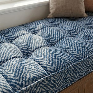May include: A window seat cushion with a blue and white chevron pattern. The cushion is tufted, with each tuft creating a rounded shape. A beige pillow rests in the background, near a window.