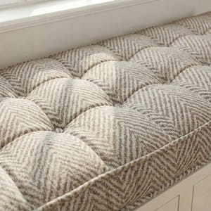 May include: A padded bench cushion in a neutral colour scheme. The cushion has a repeating pattern of rounded squares with a herringbone design in beige and grey. It is set within a white wooden frame.