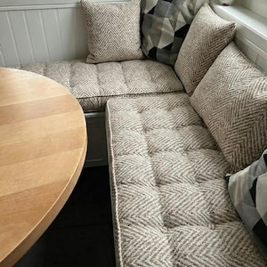 May include: A cozy corner bench with light brown and white patterned cushions and pillows. The cushions have a button-tufted design. A round wooden table is partially visible in the foreground. The pillows have a herringbone pattern.