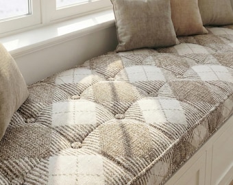 May include: A window seat cushion with a diamond pattern in shades of brown and white. The cushion is tufted with buttons and surrounded by neutral-colored throw pillows. The window frame is white.