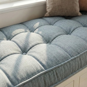 May include: A light blue, button-tufted cushion with a rectangular shape. The cushion is made of a textured fabric. It is placed on a white window seat, with a brown cushion in the background. The cushion's dimensions are approximately 60 cm x 120 cm.