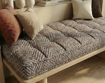 May include: A wooden bench with a tufted cushion featuring a brown and beige herringbone pattern. Several throw pillows in shades of brown and beige are arranged on the bench. Decorative vases and objects are on a shelf in the background.