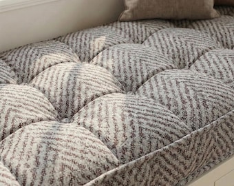 May include: A tufted window seat cushion with a geometric pattern in shades of gray and burgundy. The cushion is rectangular and has a series of rounded, padded sections. A neutral-colored pillow rests on the cushion.