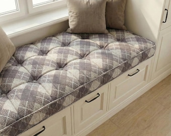 May include: A window seat with a tufted cushion featuring a gray and white plaid pattern. Two beige pillows rest on the cushion. The seat is built into a white cabinet with black handles.