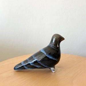 Pode incluir: Estatueta de pássaro de cerâmica com acabamento brilhante. O pássaro é principalmente preto com listras azuis horizontais no corpo. A cabeça é marrom mais escuro e está em uma superfície clara.