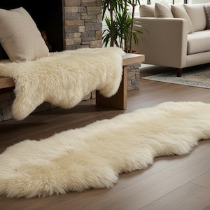 Puede incluir: Dos alfombras de piel de oveja color crema. Una está sobre un suelo de madera, la otra está sobre un banco de madera. Las alfombras tienen una textura suave y esponjosa, para un salón.