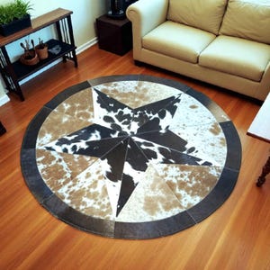 May include: A round rug with a star design. The rug features a star in the center made of black and white cowhide. The star is surrounded by sections of brown and tan cowhide, with a dark brown border. The rug is on a hardwood floor.