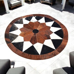 May include: A round cowhide rug with a geometric starburst design. The rug features a central black circle, surrounded by alternating brown, white, and black triangular shapes. The outer edge is a darker brown, creating a defined border. This decorative rug is suitable for indoor or outdoor use.