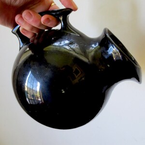 May include: A glossy black ceramic pitcher with a rounded body and a curved spout. The pitcher has two small handles and reflects the surrounding environment. The overall design is smooth and modern.