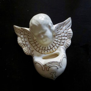 May include: A white ceramic angel figurine with gold accents. The angel has wings and is holding a heart-shaped container.
