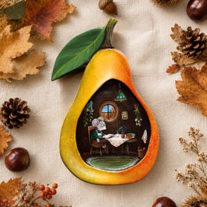 Könnte beinhalten: Ein birnenförmiges Dekorationsobjekt mit gelb-orangem Farbverlauf und einem grünen Blatt. In der Mitte befindet sich eine detaillierte Illustration einer Maus mit Brille, die an einem Tisch in einem Raum mit rundem Fenster und grünem Teppich sitzt.
