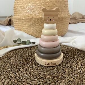 Könnte beinhalten: Ein Holz-Steckspielzeug mit einem Bärenkopf und Ringen in Weiß, Beige, Rosa und Braun. Auf der Basis ist der Name "Luisa" eingraviert. Das Spielzeug steht auf einer gewebten Matte, mit einem Weidenkorb im Hintergrund. Ein Kinderspielzeug.