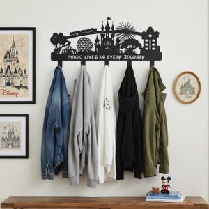 Könnte beinhalten: Schwarzer Metall-Kleiderständer mit einer Silhouette eines Schlosses, Zuges und Parksymbolen. Der Ständer hat fünf Haken und den Text "MAGIC LIVES IN EVERY JOURNEY". Verschiedene Jacken und Kapuzenpullover hängen an den Haken.