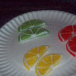 May include: A white paper plate with four slices of fruit: two green lime slices, one red orange slice, and one orange slice.