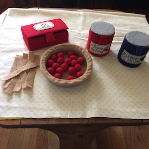 May include: A tabletop scene with felt play food for a pretend pie making activity. There is a red pie crust box, a red can labeled "Cherry Pie Filling", a blue can labeled "Blueberry Pie Filling", a bowl of red felt cherries, and a pile of tan felt pie crust strips.