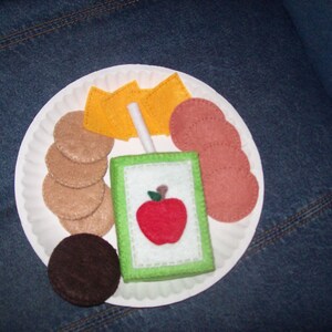May include: A white paper plate with a felt apple juice box, felt crackers, felt cheese slices, and felt bologna slices.