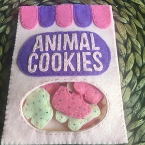 May include: A pink felt bag with a purple felt flap that says "ANIMAL COOKIES". The bag has a clear window showing pink and white felt animal cookies inside.