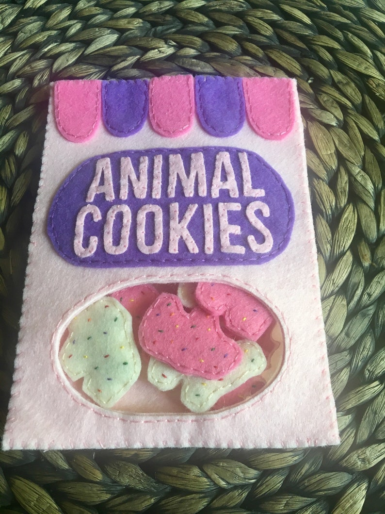 May include: A pink felt bag with a purple felt flap that says "ANIMAL COOKIES". The bag has a clear window showing pink and white felt animal cookies inside.