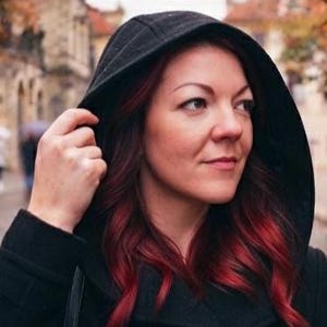 May include: A black hooded coat worn with the hood pulled up over the head. The garment features a deep black fabric construction with a structured hood that frames the face, showcasing long, wavy hair with vibrant red highlights and dark roots.