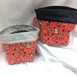 May include: Two fabric storage containers. The containers are made of orange fabric with a repeating pattern of dancing cat figures. The top edges are lined with gray fabric. One container has a black top edge and a red drawstring.