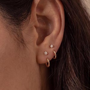 May include: A set of gold-toned jewelry worn in the ear, consisting of two small hoop earrings and two stud earrings featuring clear, round-cut stones. The jewelry is arranged in a multi-piercing configuration along the earlobe and cartilage.