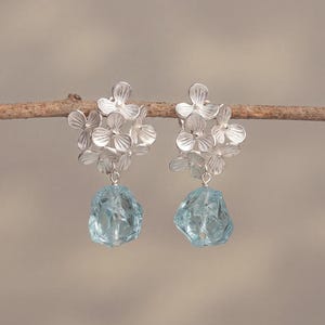 Könnte beinhalten: Silberne Ohrringe mit floralen Details. Jedes Ohrring-Design zeigt eine Ansammlung von blütenförmigen Elementen und einen hellblauen Edelstein. Die Ohrringe hängen an einem dünnen, braunen Ast vor einem neutralen Hintergrund.